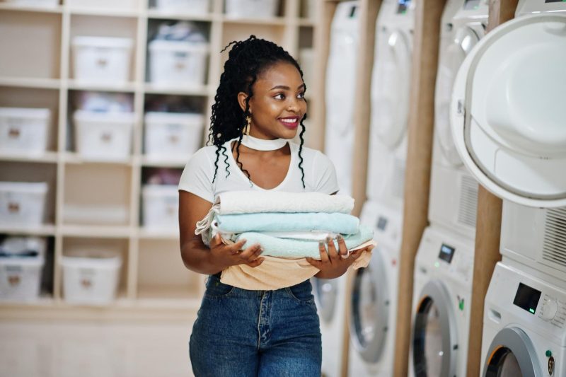 cheerful-african-american-woman-with-towels-hands-near-washing-machine-selfservice-laundry (1)